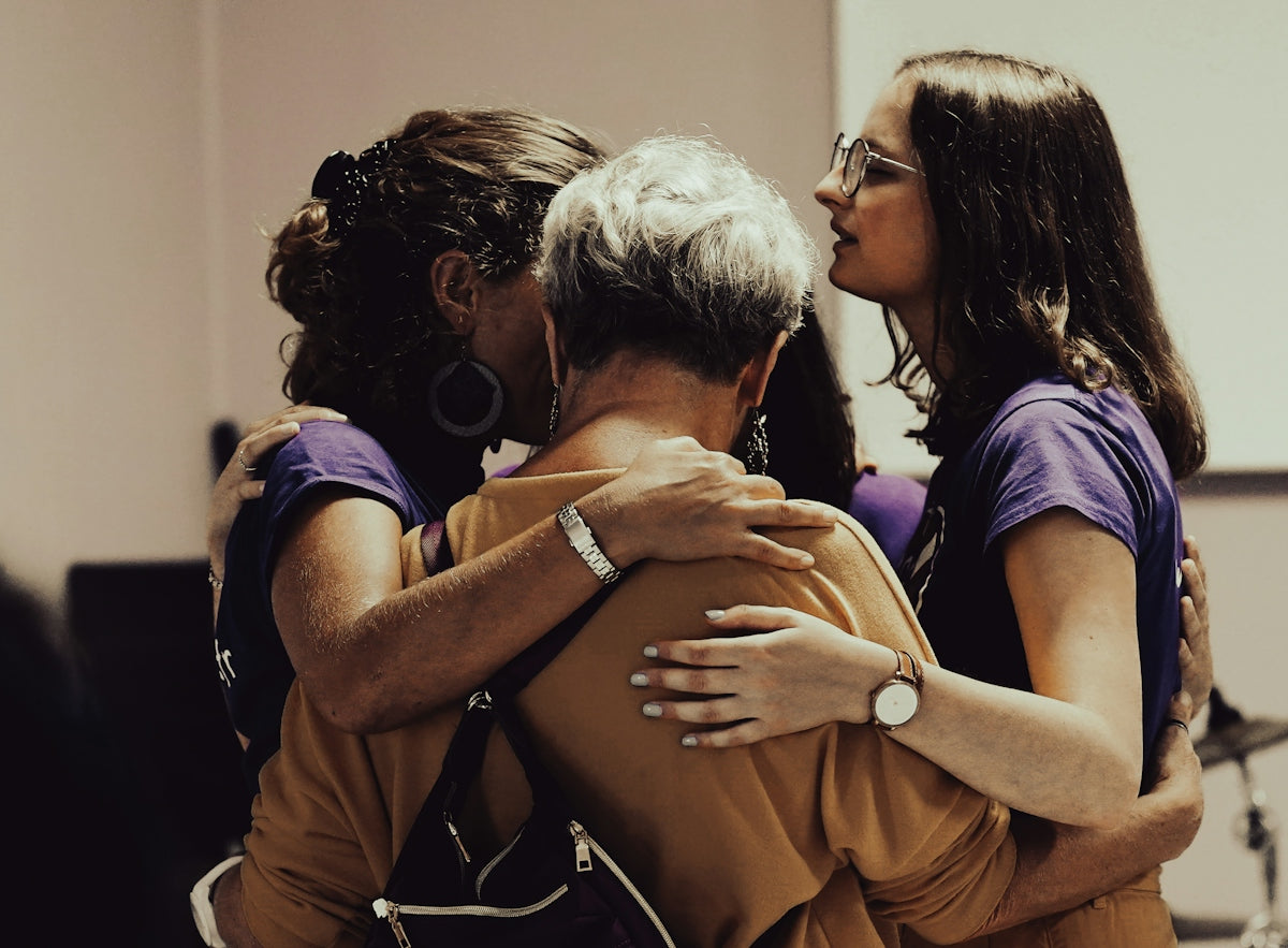 Three women in a group hug, two wearing purple shirts and one in mustard