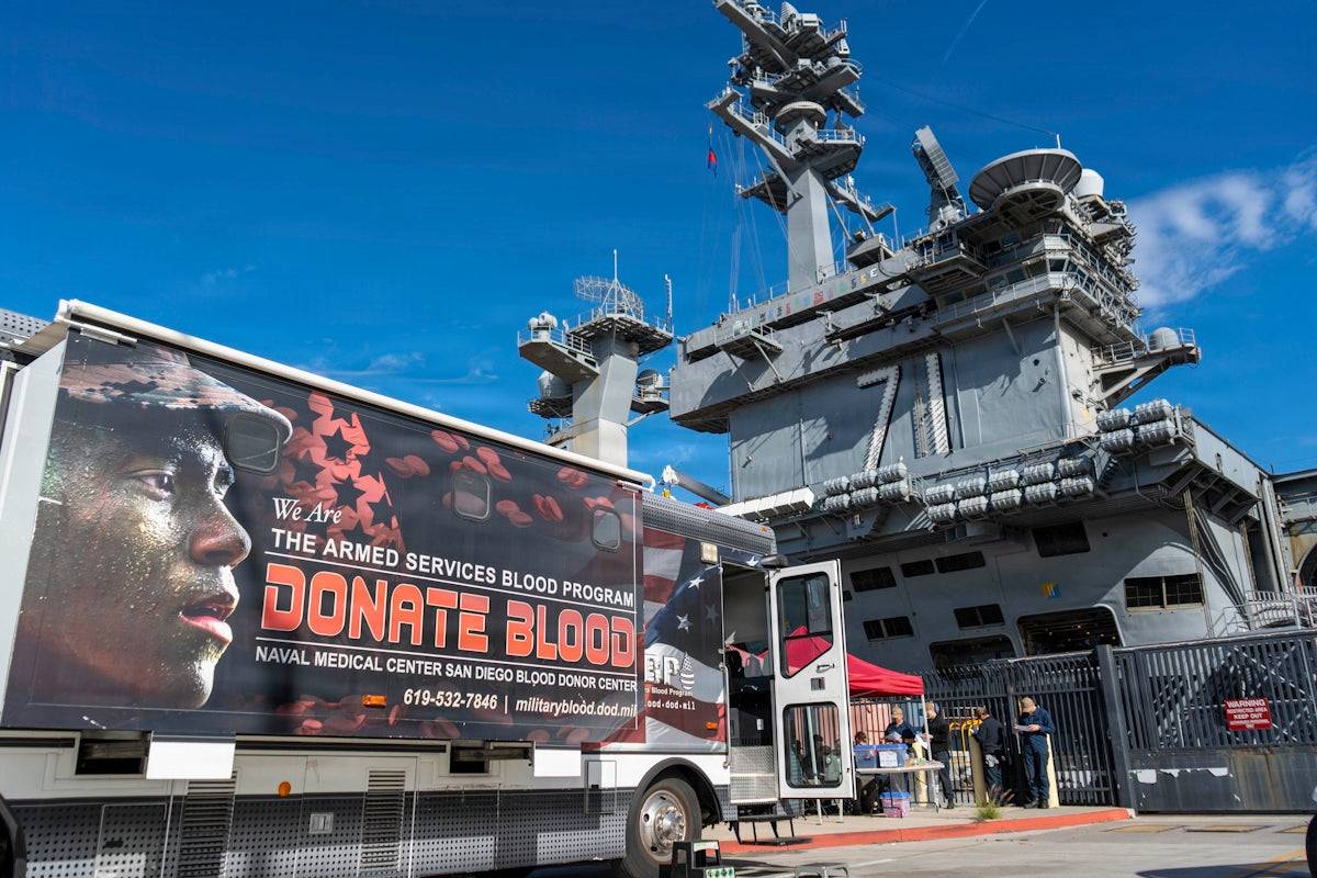 Blood donation truck parked near a large aircraft carrier.