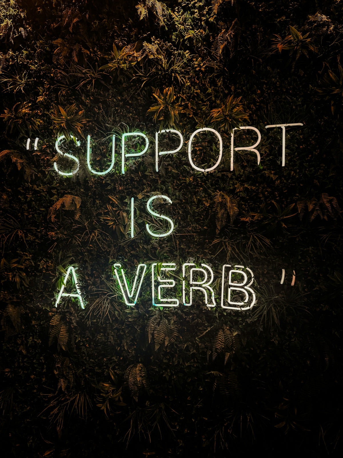 A neon sign reading SUPPORT IS A VERB on a dark green leafy wall