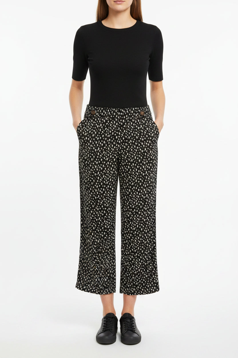 A woman wears black tapered trousers with a white speckled print and button details at the hem.
