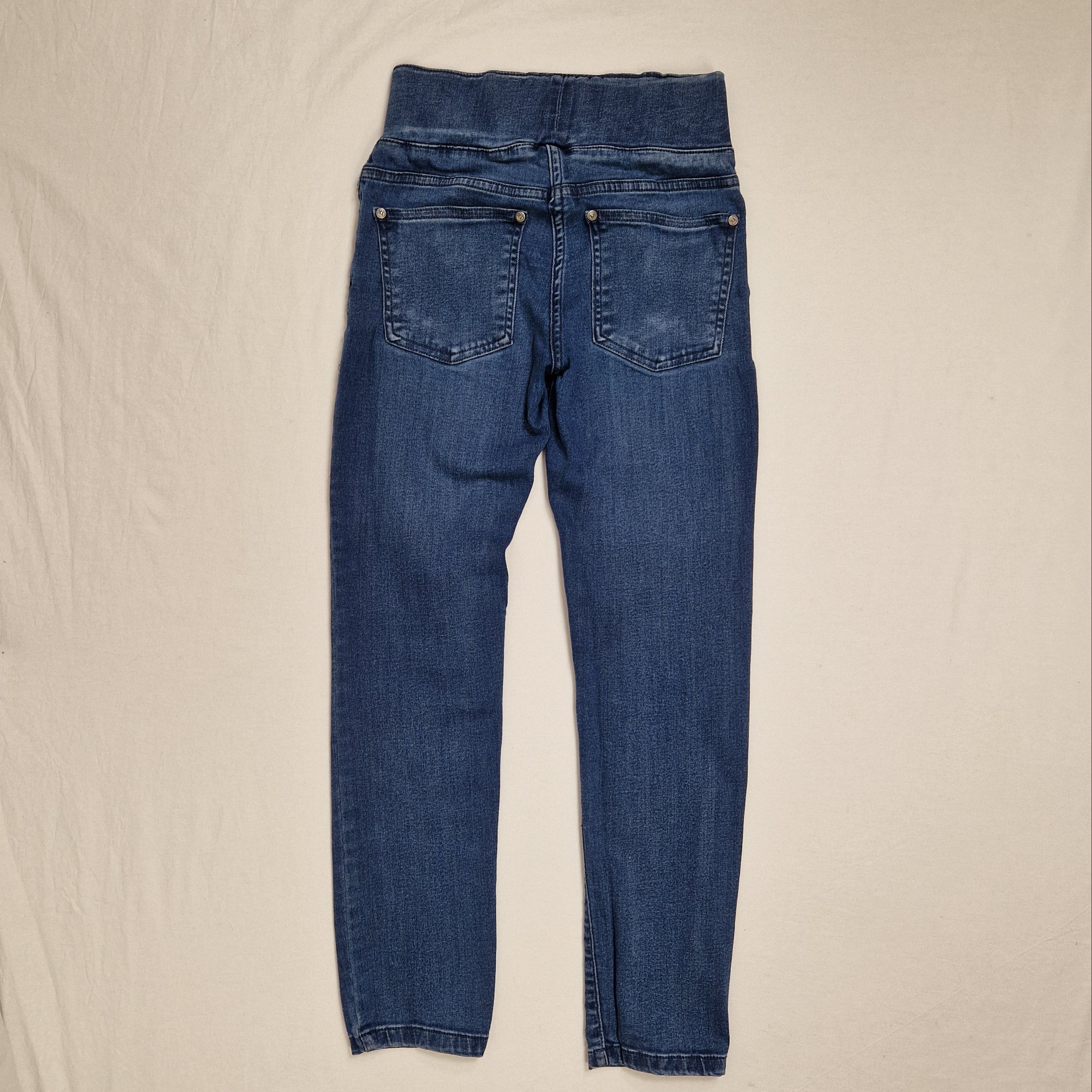 A pair of blue denim jeans laid flat on a light beige fabric background, showing the back pockets and waistband details.
