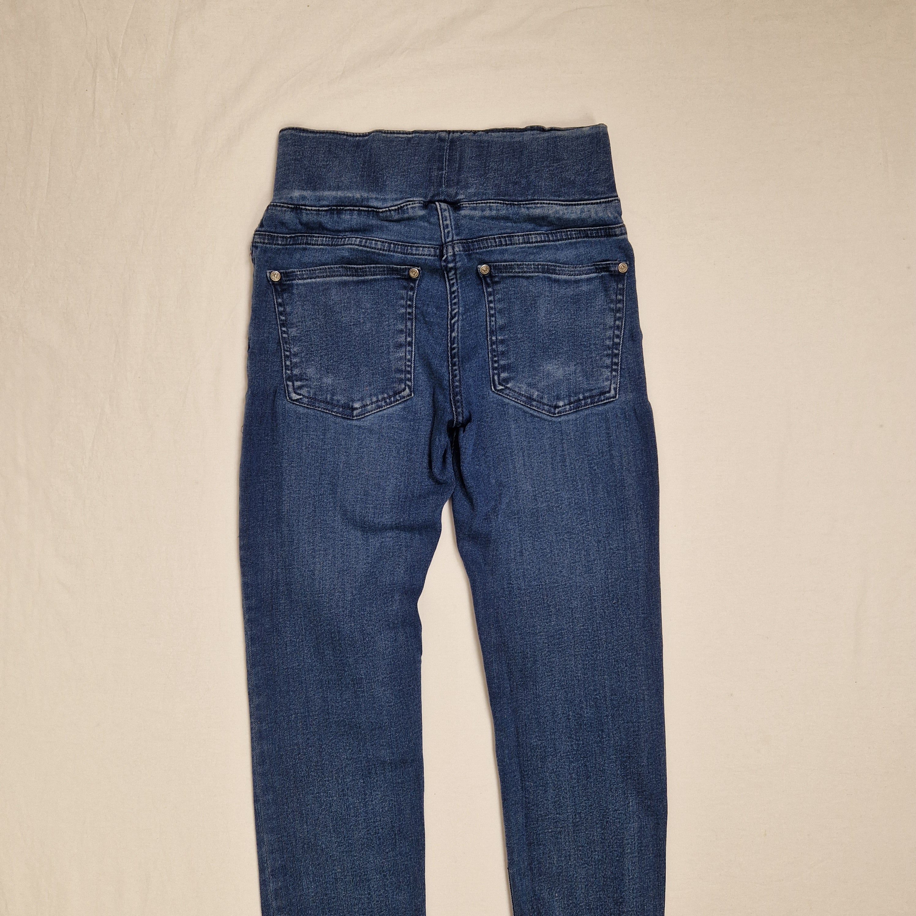 A pair of blue denim jeans laid flat on a light beige fabric background, showing the back pockets and waistband details.