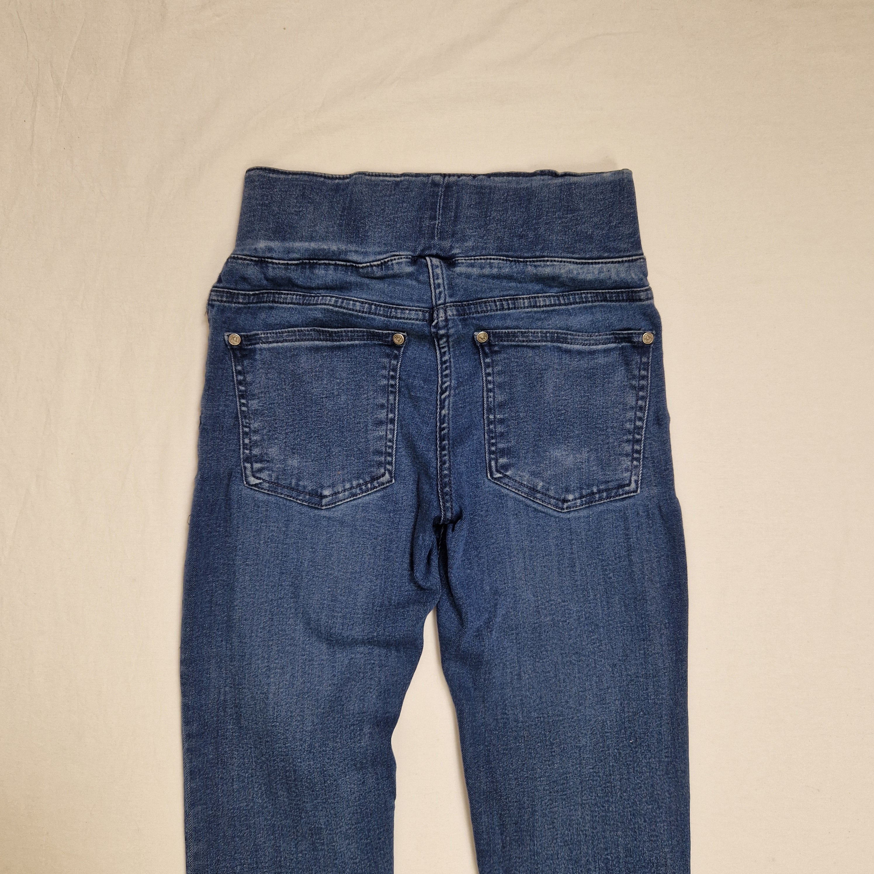 A pair of blue denim jeans laid flat on a light beige fabric background, showing the back waistband, belt loops, and rear pockets.