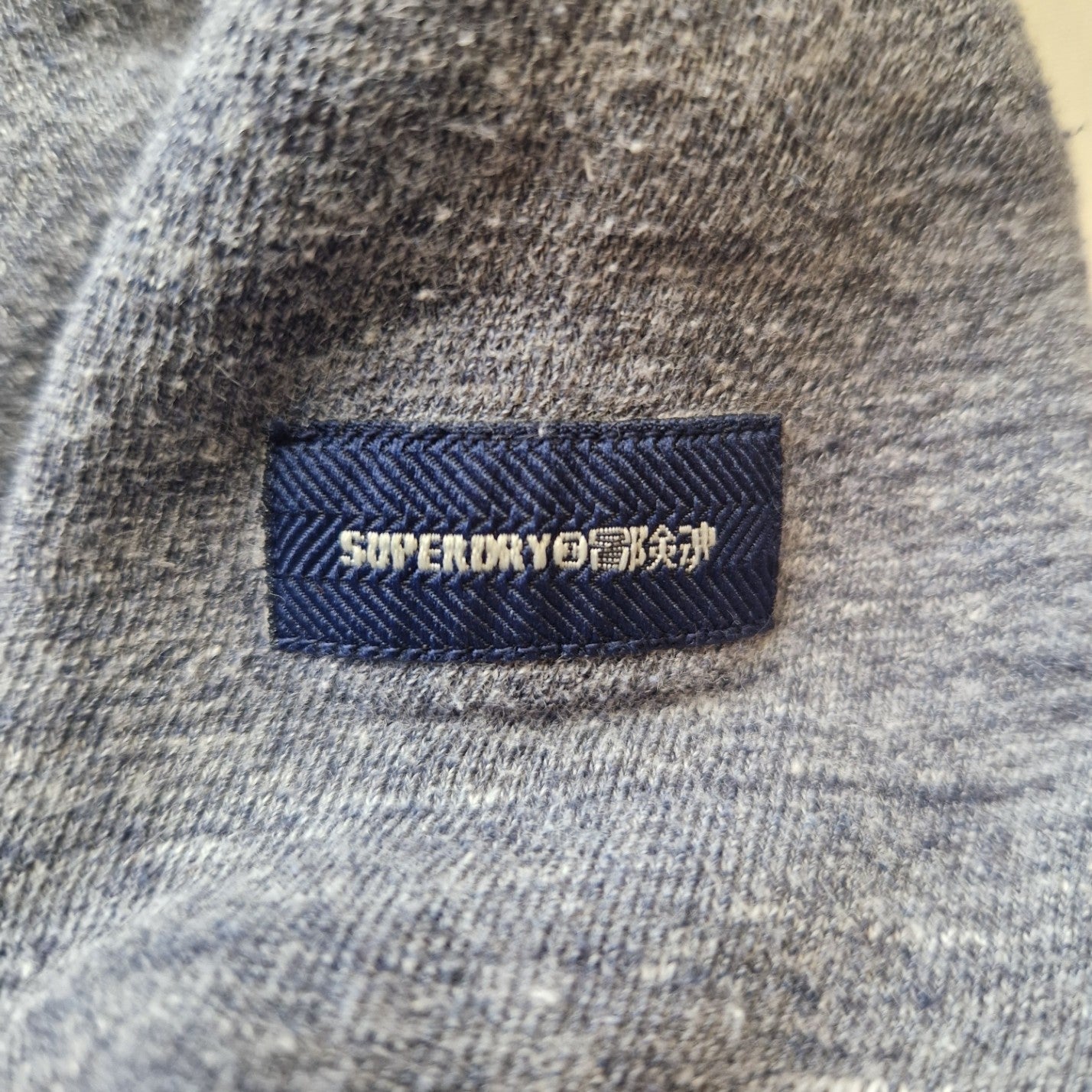 A close-up of a grey Superdry pullover hoodie featuring a blue rectangular brand logo patch.