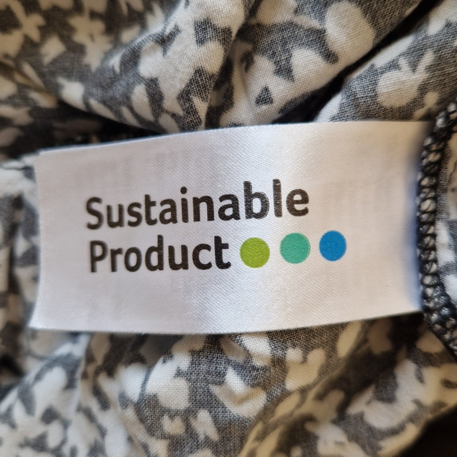 A white clothing tag with the text Sustainable Product and three colored dots on a floral dress.